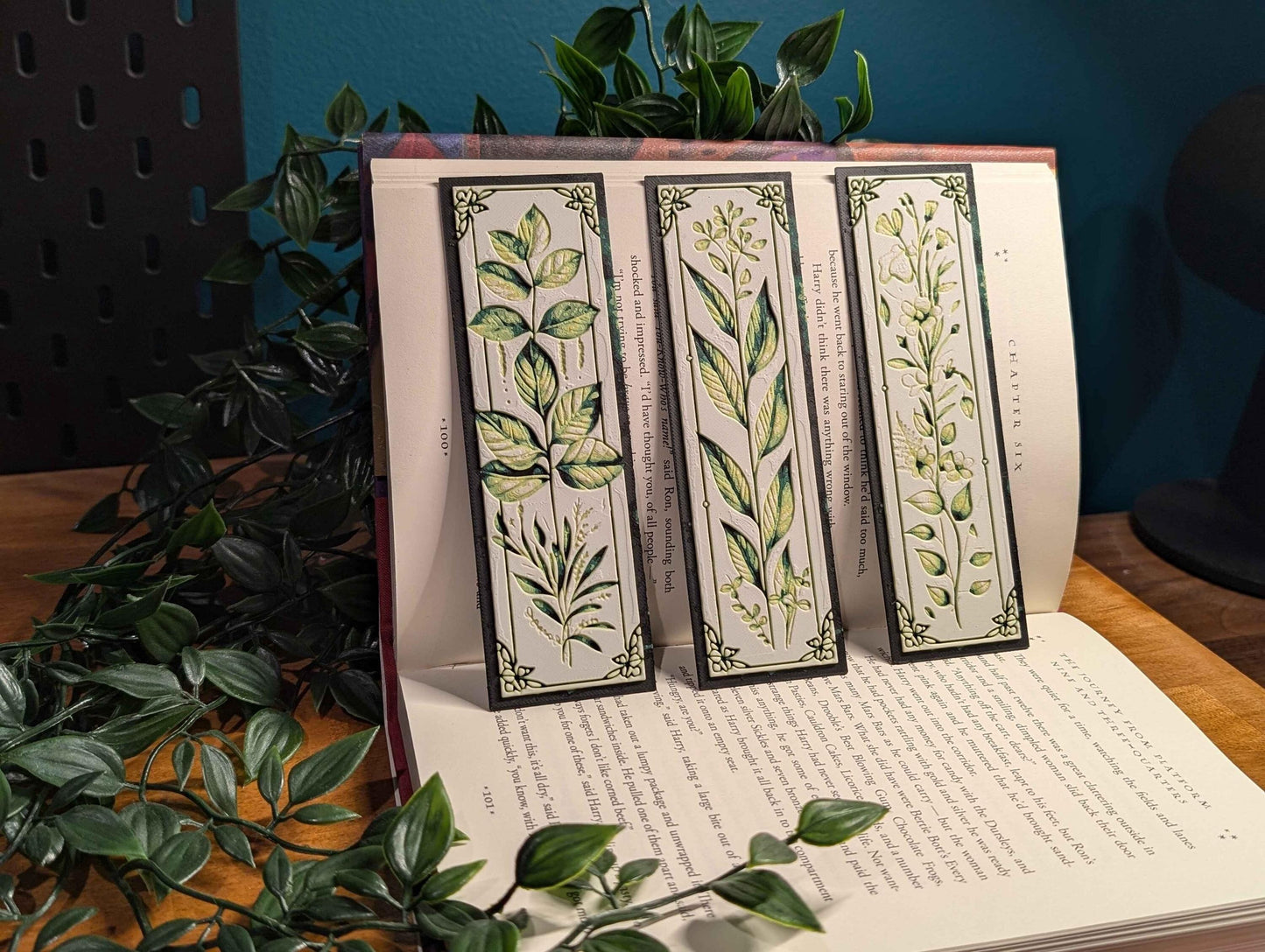 May include: Three botanical bookmarks with dark gray borders and cream-colored backgrounds. Each bookmark features a different illustration of stylized green and yellow plants and a short excerpt of text. The bookmarks are displayed against a book, partially visible, with text. The overall theme is literary and nature-inspired.