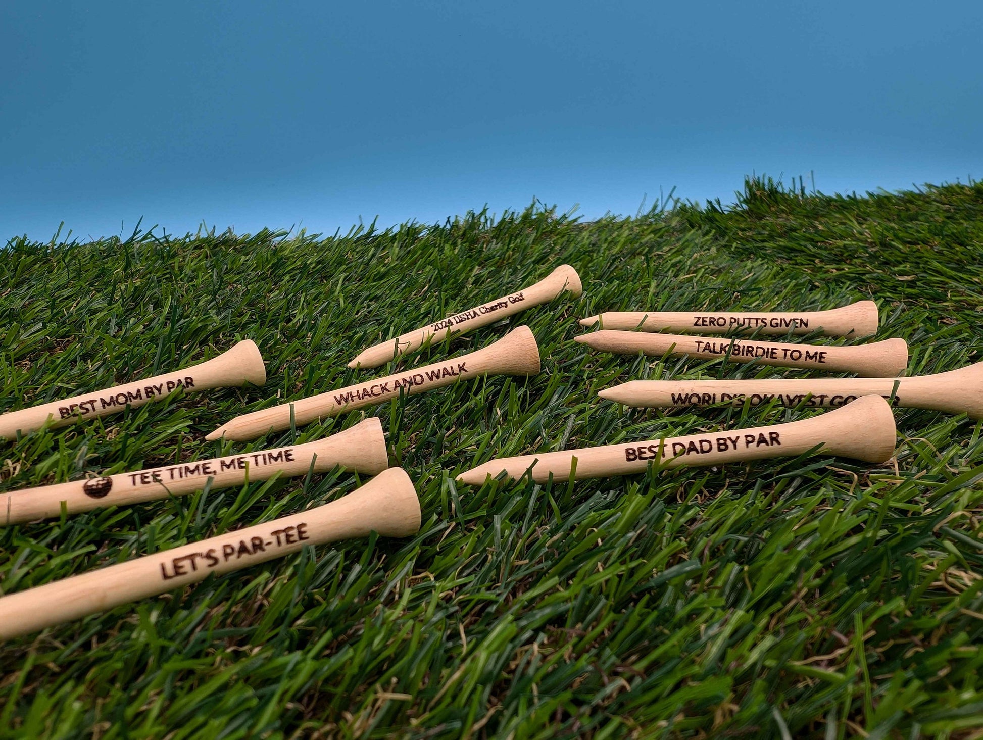 May include: A collection of eight wooden golf tees with various engraved sayings, including &#39;Best Mom by Par&#39;, &#39;Tee Time, Me Time&#39;, &#39;Let&#39;s Par-Tee&#39;, &#39;Whack and Walk&#39;, &#39;Zero Putts Given&#39;, &#39;Talk Birdie to Me&#39;, &#39;World&#39;s Okayest Golfer&#39;, and &#39;Best Dad by Par&#39;. The tees are displayed on a bed of green artificial grass.