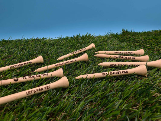 May include: A collection of eight wooden golf tees with various engraved sayings, including &#39;Best Mom by Par&#39;, &#39;Tee Time, Me Time&#39;, &#39;Let&#39;s Par-Tee&#39;, &#39;Whack and Walk&#39;, &#39;Zero Putts Given&#39;, &#39;Talk Birdie to Me&#39;, &#39;World&#39;s Okayest Golfer&#39;, and &#39;Best Dad by Par&#39;. The tees are displayed on a bed of green artificial grass.